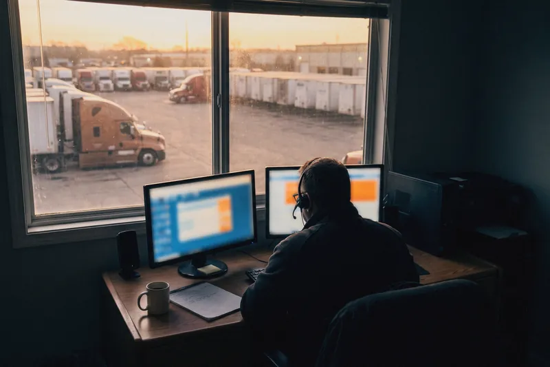 A freight broker on the phone at a desk overlooking a truck yard at dawn
